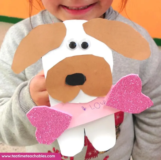 Easy Valentine craft for kids featuring a paper puppy with googly eyes and brown ears, holding pink glitter hearts and displaying "LOVE." Classroom setting.