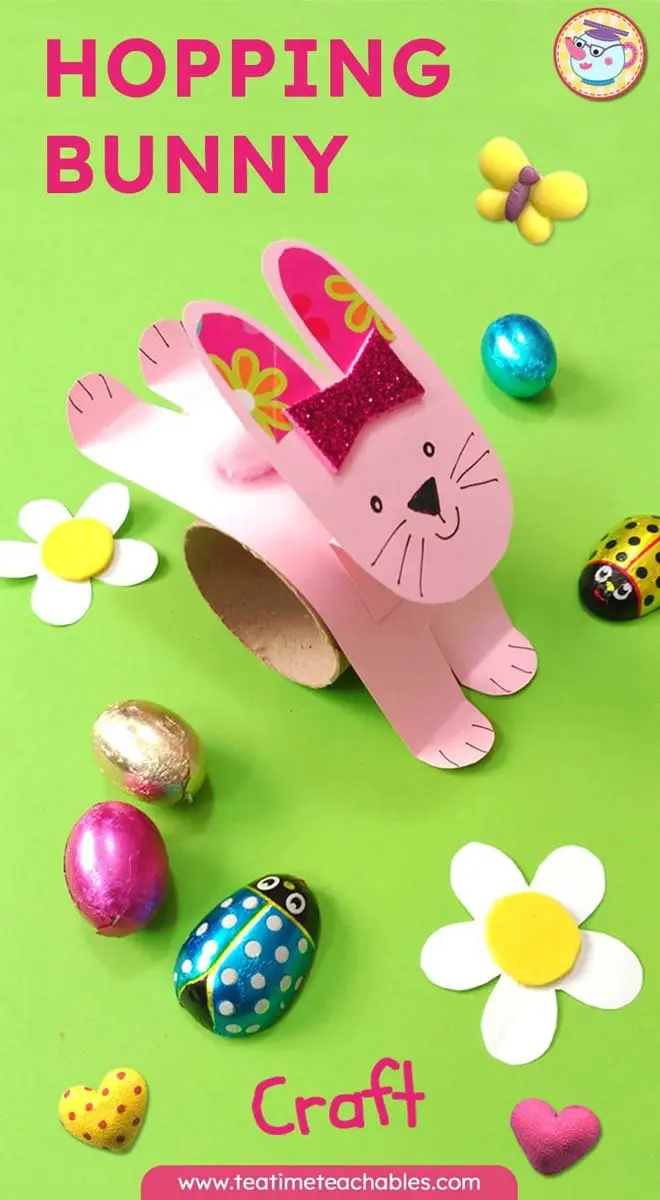 A cute pink bunny rabbit craft for kids made from paper, with a glittery bow, stands on a green background. Surrounding it are colorful Easter eggs, a daisy, and a toy ladybug.