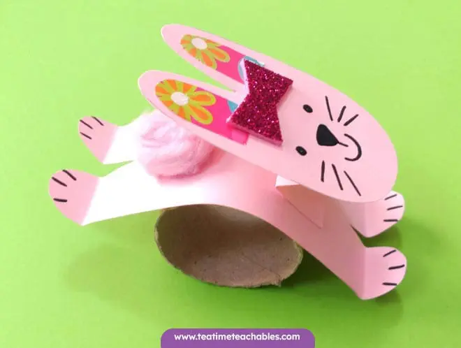 A cute pink bunny rabbit craft for kids made from paper, with a glittery bow, stands on a green background. 