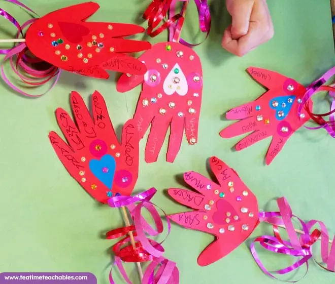 Lots of childrens' red handprint crafts decorated with hearts and ribbons on a table. Easy Valentine's Day craft for kids.
