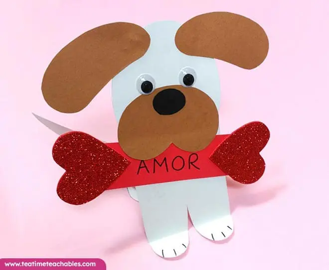 Manualidad fácil de San Valentín para niños: perrito de papel con ojos móviles y orejas marrones, sosteniendo un corazón rojo y la palabra "AMOR". Fondo rosa, estilo divertido.