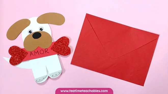 Manualidad fácil de San Valentín para niños: perrito de papel con ojos móviles y orejas marrones, sosteniendo un corazón rojo y la palabra "AMOR". Con un sobre rojo.